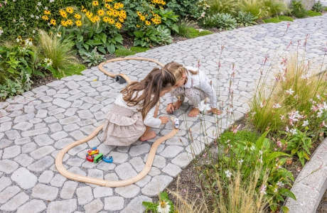 Zwei Mädchen spielen auf einem Gehweg aus kleinen, rundlichen Pflastersteinen GDM.Bolero stone mit Holzfahrzeugen