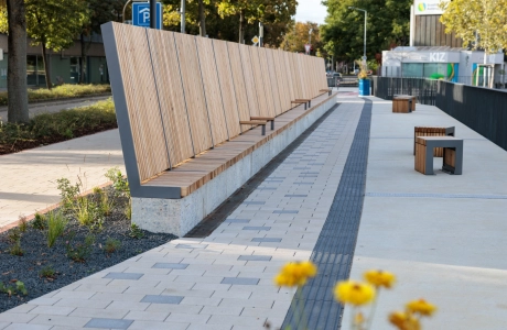 Promenade with long wooden seat backs and individual stools, laid out along a path paved with beige GDM.KLIMASTEIN; in the background trees, street area and a modern building.