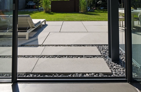 großformatige hellgraue Terrassenplatten und Sonnenliege hinter Schiebetür aus Glas mit schwarzem Türrahmen