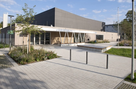 Modern exterior façade of a sports hall with anthracite-coloured panels in the upper area and wooden cladding in the lower area. In front of this is a forecourt paved with lime-grey, permeable concrete blocks with shrubs, grasses and a small tree.