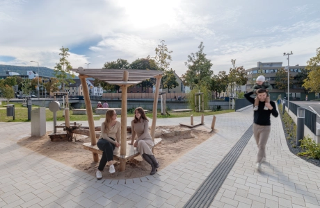 Outdoor area with a small wooden playground and neighbouring green areas; two people sit at the edge of the sand play area while another person walks past with a child. The surrounding path is paved with concrete blocks and leads through the park-like grounds.