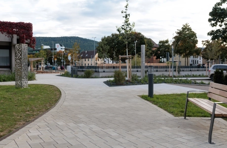 Curved path made of light-coloured concrete block, embedded in a well-tended outdoor area with young trees, flowerbeds and lawns. A wooden bench stands at the edge, next to it are further planting areas and design elements.