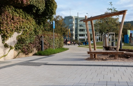 Outdoor area with a curved path made of beige GDM.KLIMASTEIN, bordering a green wall and a sand play area with wooden elements. In the background are other areas of the park with paths, plants and street furniture.