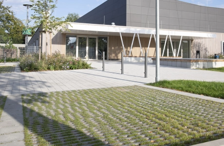 In the foreground is a car park made of grass paving stones. Behind it is a path - paved with permeable climate stone - and a modern sports hall with an anthracite-coloured façade and wooden cladding. Surrounding it are green trees and shrubs.