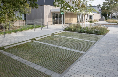 Close-up of a car park made of green grass paving stones. The individual parking spaces are separated by grey concrete blocks. Towards the front, the parking spaces are bordered by grey concrete elements. The wooden façade of the modern sports hall and anthracite-coloured bicycle racks can be seen in the background. The paths around the car park are paved with GDM.KLIMASTEIN.