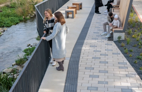 Promenade along a stream with seating and a path made of light-coloured GDM.KLIMASTEIn, into which grey paving stones are regularly integrated. Several people are standing along the railing, while others are sitting on the wooden benches in the background.