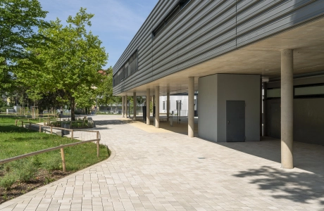 The open schoolyard is surrounded by green meadows, trees and seating elements. The ecological paving, laid out in a wild formation, features beige concrete blocks in different formats. The school building on the right-hand side of the picture stands on concrete pillars and is clad in dark gray.