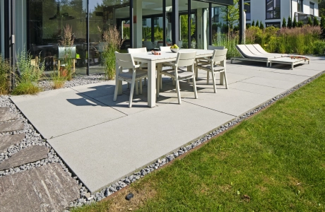 heller Esstisch mit Stühlen auf großformatigen hellgrauen Terrassenplatten und Rasen vor Glasfront eines modernen Hauses