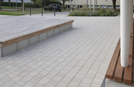 Close-up of lime-grey, permeable concrete blocks. Next to it is a concrete bench with wooden slats as a seat. Several white supports end in it. On the other side there is also a concrete bench with a wooden seat.