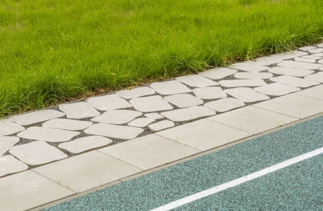 Ein mit rundlichen Betonsteinen gepflasterter Weg, welcher eine petrolfarbene Sportfläche von einer grünen Wiese trennt.