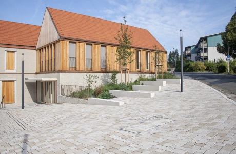 Modern building with wooden façade and tiled roof next to an ecological surface covering with benches.