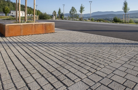 Nahaufnahme von Naturbeige schattiertem Betonpflaster. Im Hintergrund ist eine geteerte Straße und eine rostige Corthenstahleinfassung mit Holzsitzfläche um einen Baum.