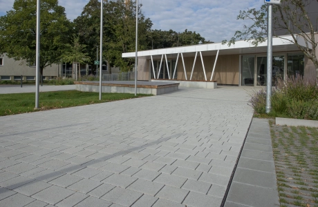 Path made of lime-grey, permeable climate stones, which leads to a sports hall. This has a white roof, which is held in place by beams running diagonally towards each other. Surrounding it are green areas with shrubs and trees.