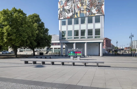 großer grau gepflasterter Platz vor schmalem Hochhaus mit hohen Fenstern und Mosaik