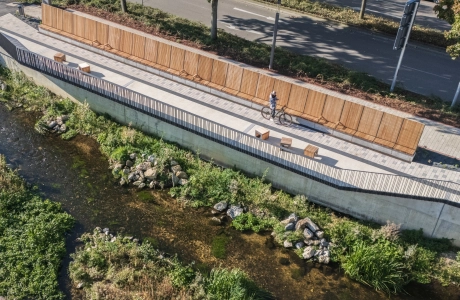 Aerial view of a riverside path along a shallow stream, lined with wooden benches and long wooden panelling. The path is paved with light-coloured concrete blocks, while a road with trees runs above it.