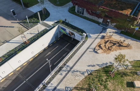 Aerial view of a modern outdoor area with a paved path made of light-coloured concrete block, adjacent to a road tunnel, a green building and a sand playground.