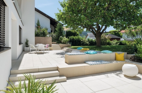 Patio slabs in a landscaped garden with a large tree and concrete seating elements.