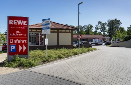 Zufahrt zum REWE Markt Christian Schulz mit Beschilderung und Fahrgasse aus GDM.KLIMASTEIN aBG, eingefasst von Grünflächen und Parkbereichen.