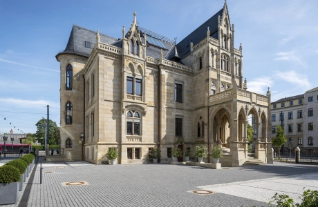 Historic villa with several storeys and elaborate architectural details. The building has a light-colored sandstone façade, ornate window frames and a distinctive entrance area with a balcony above. The paved square around the villa is well-kept and surrounded by trees and planters. Parking spaces can also be seen in the foreground