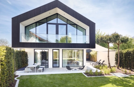 Modern two-storey house with large glass windows and a gable at the top. The building is kept in simple colors, the façade of the upper floor is dark, while the first floor is white. In the foreground is a well-tended garden with a green lawn and a terrace with garden furniture. Two slender trees and green hedges frame the outdoor area.