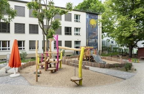 Außenbereich der Kita Wolkenschaf mit Sandkasten, Spielgeräten aus Naturholz und dem Kita-Gebäude im Hintergrund. Pflastereinfassung aus GDM.BOLERO.