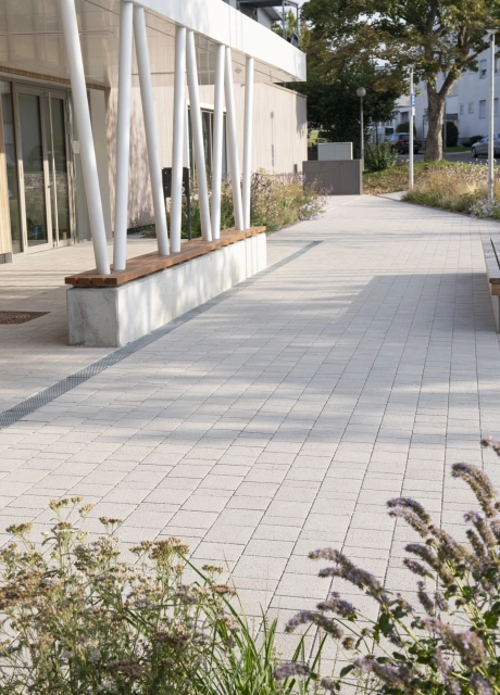 Forecourt of a modern building with a covered entrance area. The roofing is held up by white supports that run diagonally towards each other and end in concrete blocks with a wooden seat. Next to it is a path paved with lime-grey, permeable climate stones. There are flowering shrubs in the foreground.