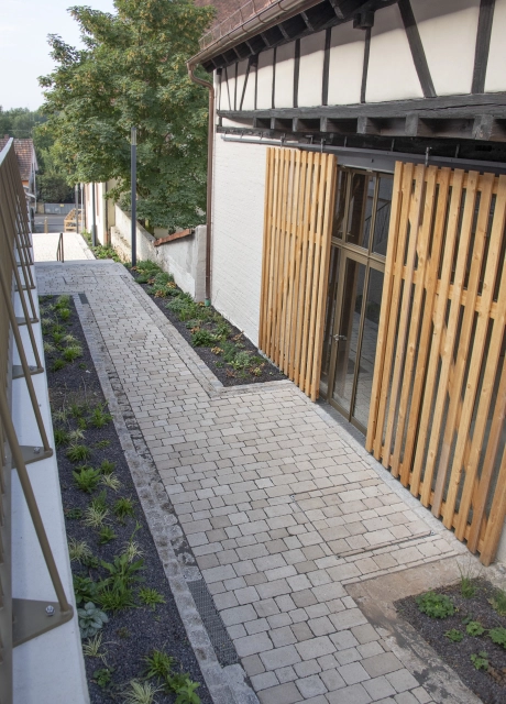 A paved path alongside a timber-framed building with wooden elements. A white wall with wooden louvres in front of large windows is visible on the right and a narrow flower bed with young plants on the left. In the background are large trees and another building.