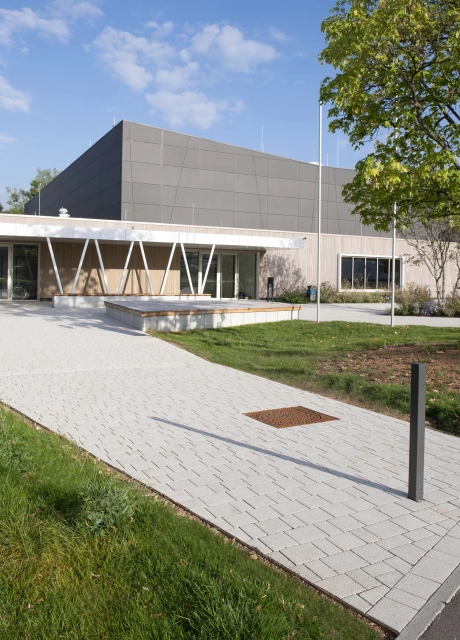 Path made of GDM.KLIMASTEIN lime-grey, permeable concrete block, which leads to a sports hall. This has a façade with anthracite-coloured panels in the upper area and wooden cladding in the lower area. There are green areas next to the path.