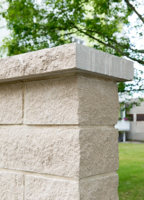 sandfarbener Abdeckstein auf gleichfarbiger Mauer vor Rasenanlage und Bäumen