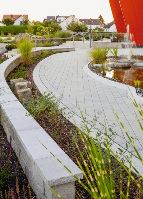 geschwungene niedrige Mauer neben hellgrau gepflasterten Gartenweg und Beetanlage mit Pflanzen