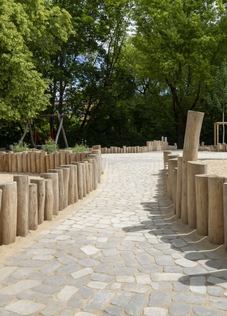 Mit GDM.BOLERO gepflasterter Weg, eingefasst von Naturstämmen. Links eine Rutsche auf Sand, rechts ein Naturholz-Spielgerät mit farbigen Akzenten.