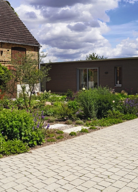 A rustic brick building with large wooden gates and a landscaped garden in the foreground. A paved path leads through the garden, which is planted with flowers and shrubs. In front of the garden is a natural beige paved courtyard laid out in rows on which a child is riding a bicycle. On the right is a modern building with large glass doors.