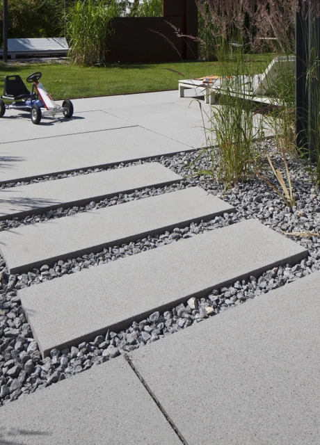 Gartenweg aus hellgrauen Platten, Splitt und Pflanzen hin zu einer Terrasse 