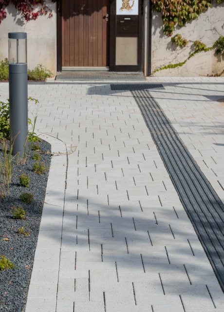 Well-kept outdoor area with a light-coloured, paved path. A continuous band of anthracite-coloured stones is integrated into the path and serves as a guiding element. To the left is a planted bed with dark-coloured grit, and to the right is a sand play area with wooden elements.