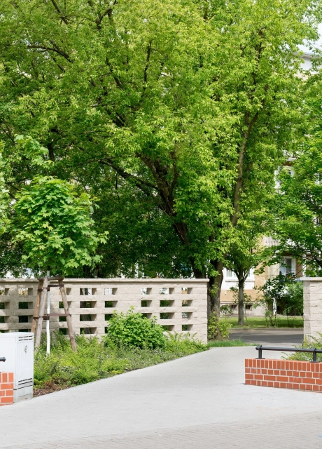 Fußgängerweg in einen Park mit Büschen und Bäumen mit Abgrenzung zweier roter kleiner und zweier sandfarbenen großen Mauern