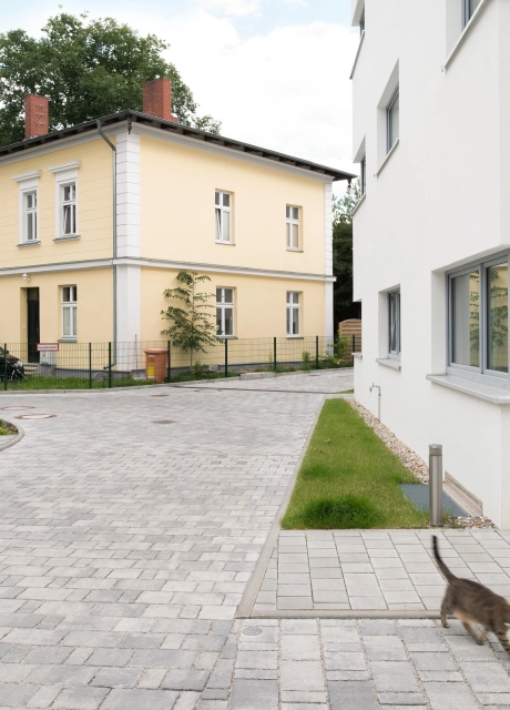 Gehweg und Einfahrtsstraße aus hellgrauem Pflaster zwischen zwei modernen weißen Gebäuden mit Blick auf orangenes Wohnhaus
