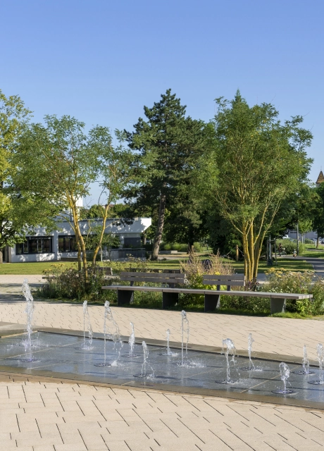 Open square in a green park environment. A water feature with fountain elements is placed in the middle of the paved square. In the background are many green areas with meadows, shrubs and trees. A building with a white façade can be seen behind it.