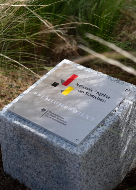 Gedenkstein mit Metallplakette in einer Gräserpflanzung. Auszeichnung als „Nationale Projekte des Städtebaus“ für ein Premiumprojekt.