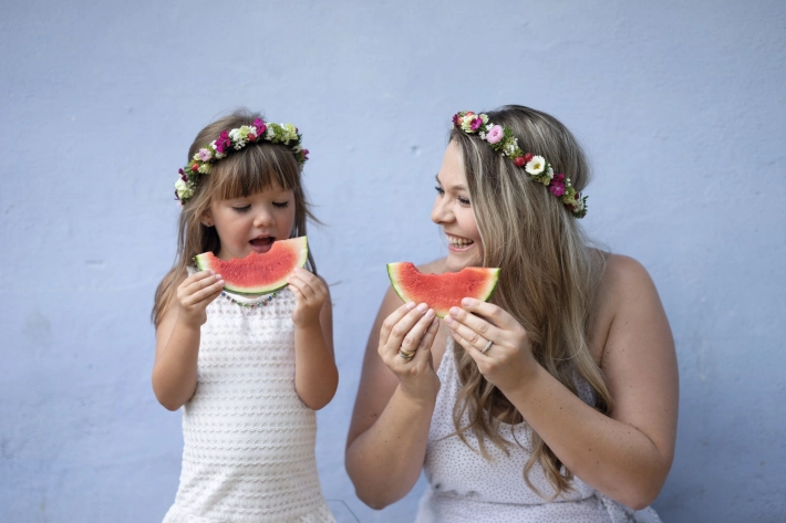 A woman and a child with wreaths of flowers in their hair eat a watermelon in front of a blue wall.