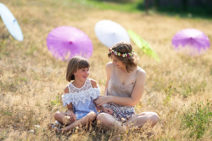 A girl and a woman sit laughing in a meadow with parasols in the background.