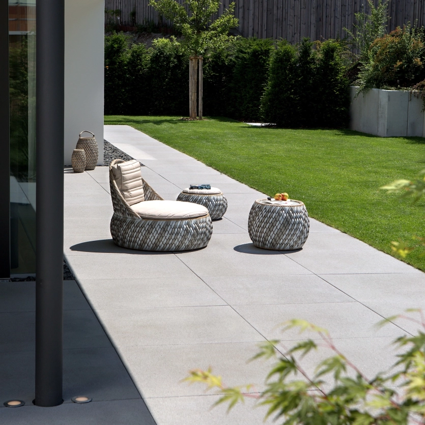 Modern terrace with large-format, gray slabs, furnished with round, woven seating furniture and a small table; bordered by a well-tended lawn and hedges in the background.