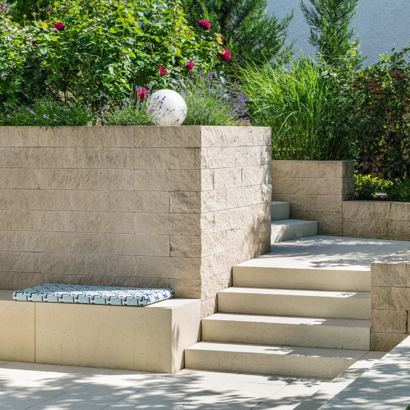 Light-colored seating, concrete walls and steps surrounded by green planted beds.