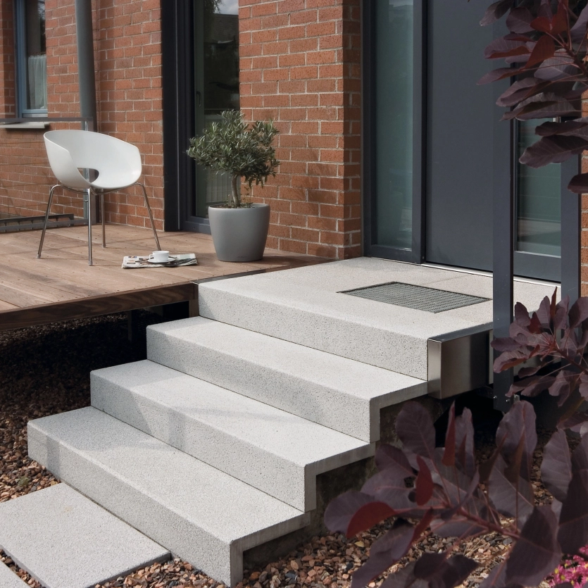 Light-colored staircase made of concrete slabs leading from a gravel-covered area to a wooden terrace; a modern white chair stands on the terrace next to a small table, in the background a brick façade with a glass door and a window.