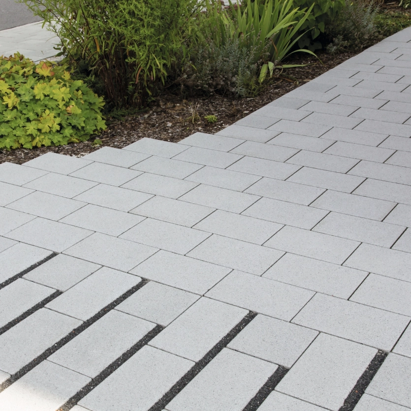 Light-colored paved walkway made of rectangular concrete slabs laid in a checkerboard pattern, adjacent to a planted garden area with shrubs and small plants.