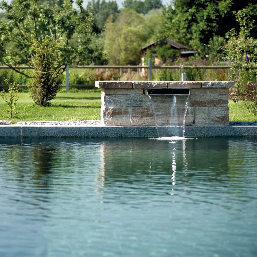 A splash shower is set into a GDM.KLASSIK wall at the edge of the pool and splashes into the water.