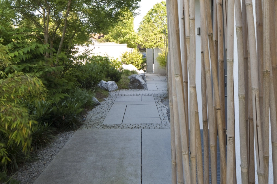 Bambusstäbe vor Gartenweg aus grauem Pflaster seitlich neben einem Wohnhaus mit Büschen und Bäumen