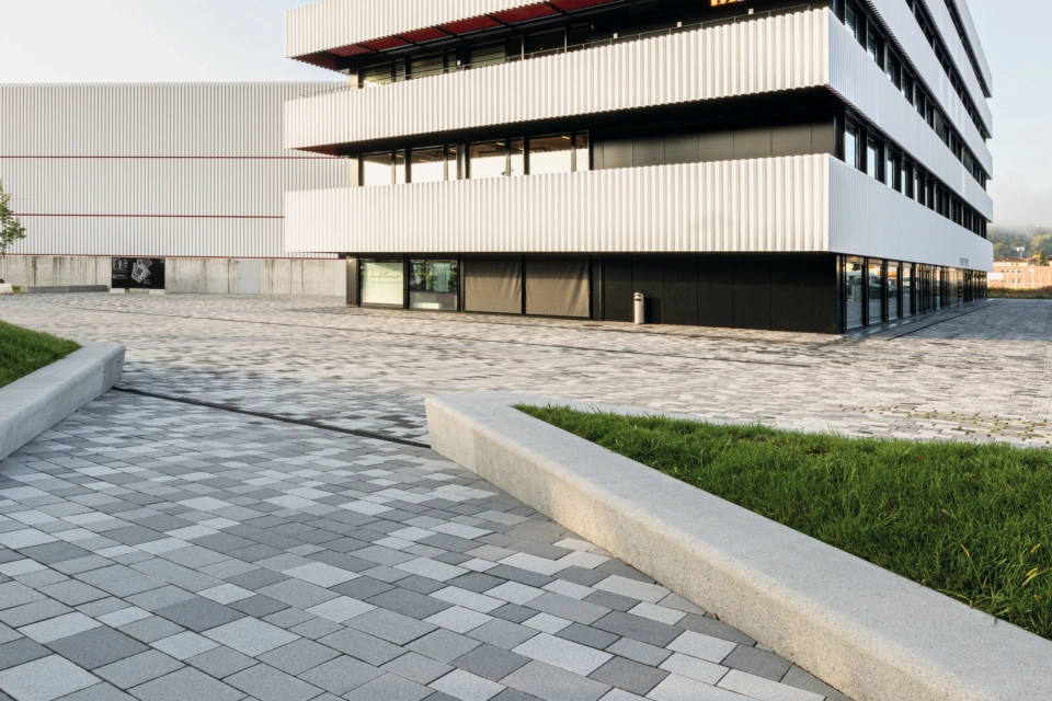 Gepflasterter Platz mit teilweise begrünter Fuge und an Grünfläche angrenzende Sitzmauern vor einem Sportzentrum