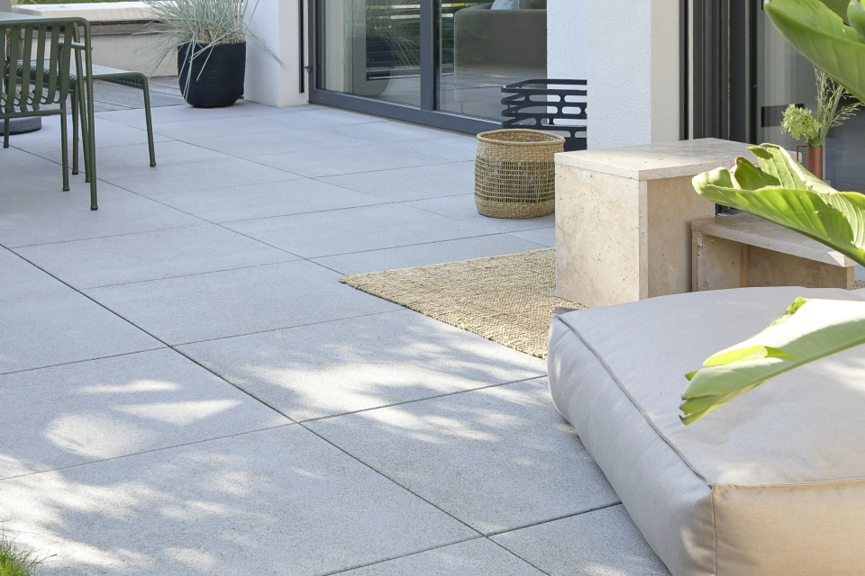 Nahaufnahme einer Terrasse mit grauen Terrassenplatten aus Beton. Dort befindet sich ein Sitzkissen, ein Tisch und Stühle. Im Hintergrund ist das Haus mit weißer Fassade und großen Fenstern.