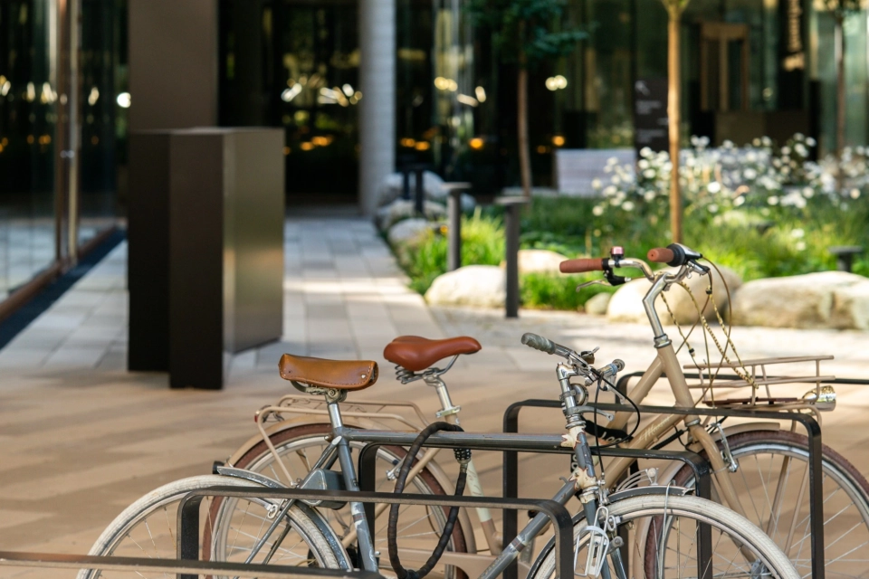 Nahaufnahme von anthrazitfarbenen Fahrradständern mit abgestellten Fahrrädern. Diese stehen auf einer mit GDM.SCENIC individual gepflasterten Fläche. Im Hintergrund ist ein großes Gebäude und grüne Beete zu erkennen.