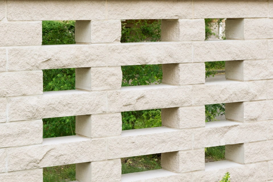 sandfarbene hohe Mauer mit rechteckigen Aussparungen durch fehlende Mauersteine vor Büschen und Bäumen